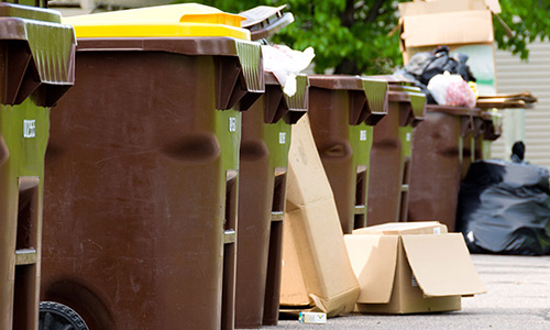 Six brown garbage cans with either brown or yellow lids on the street with cardboard boxes and garbage bags visible.