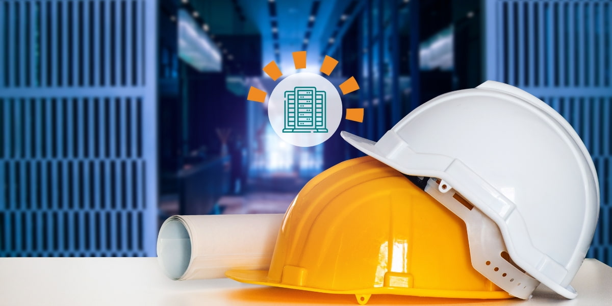 A white construction hard hat partially resting on top of a yellow construction hard hat, setting next to blue prints, next to large panels of servers..