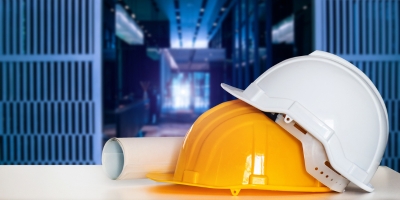 Two construction helmets, one white and one yellow, resting on top of each other and next to rolled blueprints, with a network of servers in the background.