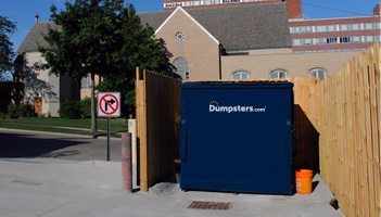 Dark blue Dumpsters.com front load dumpster in a wooden enclosure behind a building.