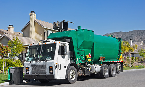 a green and white garbage truck outside of a beige house lifting up a trashcan with an automatic arm. 