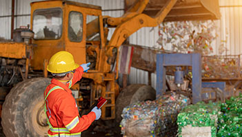 Worker in a yellow hard hat and wearing an orange, reflective suit, standing next to a working backhoe, sorting garbage at a waste facility.