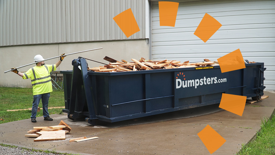 Builder with board, loading a dumpster, with two workers in background.