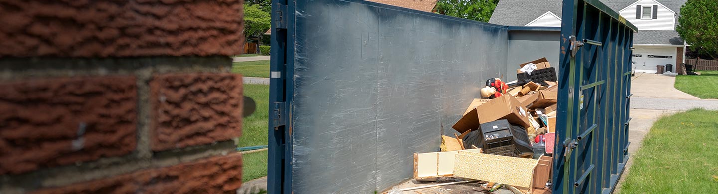 man throwing a box into a roll off dumpster in a residential driveway with a white house in the background