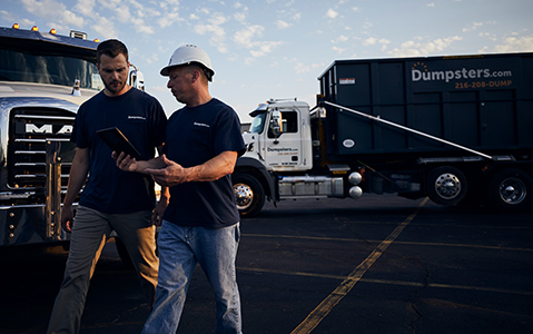 A construction worker and a driver talking over a clipboard with two Dumpsters.com dump trucks in the background.